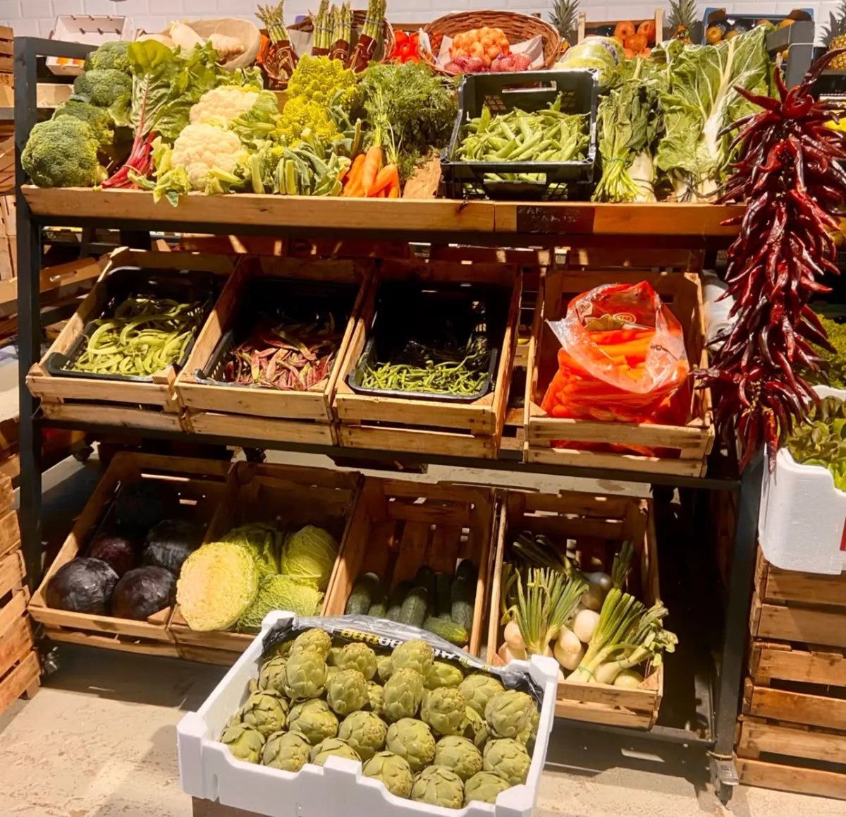 Interior de la frutería con cajas de madera llenas de verduras, alcachofas y ristras de pimientos