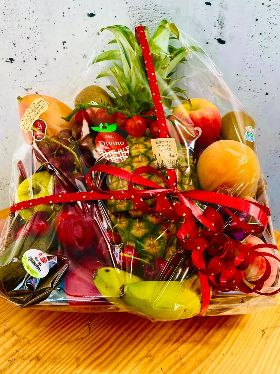 Cesta de frutas variadas con piña, uvas, plátanos y manzanas, envuelta con lazo rojo